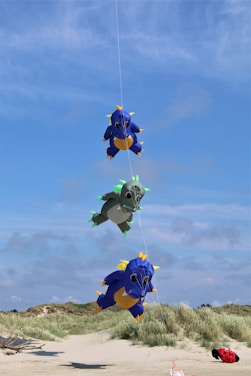 Three colorful dragon kites are tethered in a vertical line against a clear blue sky. The kites are vibrant and designed with a playful, cartoon-like appearance. In the background, there is a sandy beach with grassy dunes, adding a natural contrast to the whimsical kites.