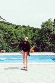 A person wearing dark, fashionable clothing and sunglasses stands on a small platform beside a swimming pool, set against a backdrop of lush green trees. The scene appears stylish and modern.