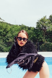 A person wearing a glamorous black fur coat and large black sunglasses is striking a dramatic pose beside a swimming pool. The person has bold red lipstick, and their hair is styled smoothly. The background features lush green trees under a bright sky.