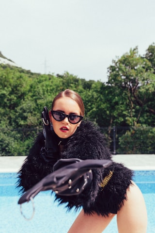 A person wearing a glamorous black fur coat and large black sunglasses is striking a dramatic pose beside a swimming pool. The person has bold red lipstick, and their hair is styled smoothly. The background features lush green trees under a bright sky.