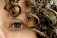 An extreme close-up of a human eye surrounded by curly brown hair. The eye is blue and the lashes are visible, while the curls frame the eye in a dynamic pattern, showcasing the texture of the hair.