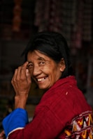 A woman is smiling warmly, her hand slightly raised close to her face. She is wearing a red garment with intricate patterns and has dark hair.