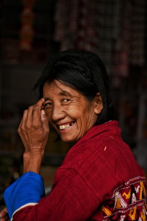 A woman is smiling warmly, her hand slightly raised close to her face. She is wearing a red garment with intricate patterns and has dark hair.