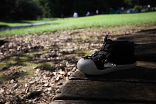 A single child's black shoe with a white toe rests on a wooden surface with dried leaves scattered around. In the background, a lush green grass field can be seen, with people blurred in the distance, suggesting a park or outdoor setting.