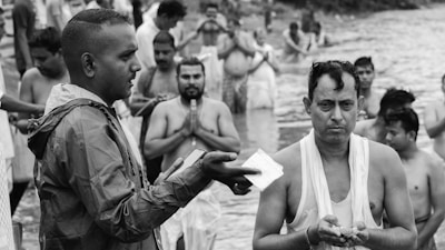 A group of people is gathered in a body of water, participating in a ritual or ceremony. One person in a raincoat is speaking or offering something to another person holding items in their hands. Others in the background are standing in water, some with hands folded in prayer.