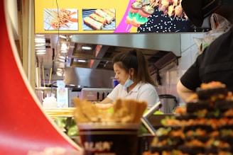 A woman is working in a kitchen or food preparation area, surrounded by various food items and cooking utensils. She is wearing a white shirt and has her hair tied back. There is a toasting machine nearby and skewer dishes displayed on a board above.