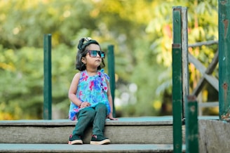 A young child wearing colorful sunglasses, a blue floral top, teal pants, and black shoes with pink accents is sitting on a wooden structure. The background is filled with lush green foliage that creates a serene and natural setting.