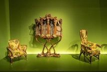 Antique furniture pieces consisting of two ornate armchairs and a decorative cabinet. The furniture is upholstered in yellow fabric with green and gray floral patterns, set against a vibrant green background. The armchairs feature curved wooden frames and the cabinet has intricate carvings and a mirrored surface.