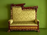 An ornate, antique wooden sofa with intricate carvings and a plush, patterned upholstery in a light green color. The wooden frame is decorated with floral motifs and features elegant curves and detailing. The background is a solid matching green, complementing the upholstery.