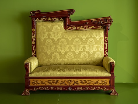 An ornate, antique wooden sofa with intricate carvings and a plush, patterned upholstery in a light green color. The wooden frame is decorated with floral motifs and features elegant curves and detailing. The background is a solid matching green, complementing the upholstery.