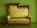 An ornate, antique wooden sofa with intricate carvings and a plush, patterned upholstery in a light green color. The wooden frame is decorated with floral motifs and features elegant curves and detailing. The background is a solid matching green, complementing the upholstery.