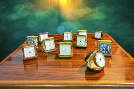 Multiple small clocks with square and circular faces are arranged on a wooden surface. The clocks have various colored faces and frames, ranging from gold to dark blue, with gold being the most predominant. A soft green bokeh background adds depth, with a warm light source above highlighting the scene.