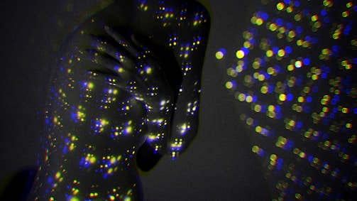 A person is enveloped in darkness, illuminated by multicolored light dots projected onto their skin, creating a cosmic and abstract atmosphere. The multitude of small, bright circles cover the person's figure, blending with the subtle shadows of the background.