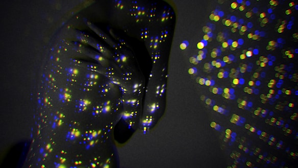 A person is enveloped in darkness, illuminated by multicolored light dots projected onto their skin, creating a cosmic and abstract atmosphere. The multitude of small, bright circles cover the person's figure, blending with the subtle shadows of the background.