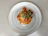 A plate of spaghetti topped with tomato sauce, cherry tomatoes, grated cheese, and fresh basil garnish is placed on a simple, light gray plate against a neutral colored background.