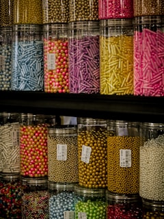 Colorful jars filled with a variety of small decorative sprinkles, including pearls and rods, arranged tightly on shelves. The jars are transparent, allowing the vibrant colors of the contents to stand out against the darker background.