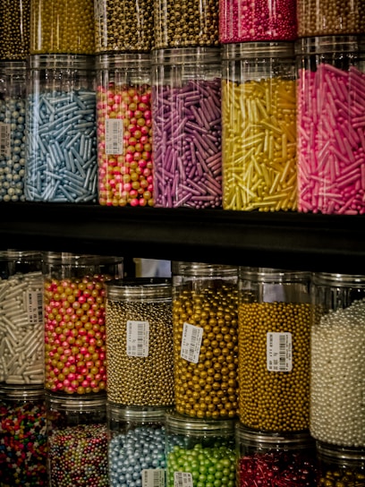 Colorful jars filled with a variety of small decorative sprinkles, including pearls and rods, arranged tightly on shelves. The jars are transparent, allowing the vibrant colors of the contents to stand out against the darker background.