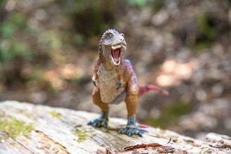 A toy dinosaur model with an open mouth stands on a textured rock, surrounded by a blurred natural background.