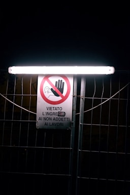 A warning sign with a prohibitory symbol, indicating restricted access to unauthorized personnel. The sign is illuminated by a bright light tube above, mounted on a wire fence. The text is in Italian, featuring a graphic of a hand and a hard hat.