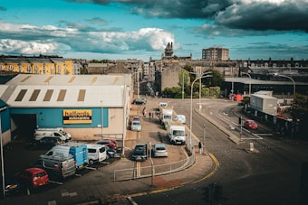 An urban scene featuring a street intersection with a mix of light and medium traffic, including cars and delivery vans. A Halfords autocentre with blue walls is prominent on the left, adjacent to small, older stone buildings. The sky is filled with large, dark clouds suggesting an overcast day.