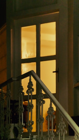 A warmly lit entrance is visible through a window with multiple panes. The soft glow suggests a cozy interior. A decorative metal railing is prominently featured in the foreground, leading towards the door.