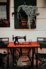 A vintage sewing machine is placed on a wooden table against a rustic wall. Above the machine, a sign with text in two languages is partially covered by green foliage. On either side of the table are metal chairs, and a lace-curtained window is partially visible to the left.
