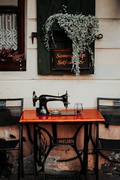 A vintage sewing machine is placed on a wooden table against a rustic wall. Above the machine, a sign with text in two languages is partially covered by green foliage. On either side of the table are metal chairs, and a lace-curtained window is partially visible to the left.
