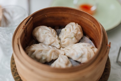 A bamboo steamer filled with freshly cooked dumplings, arranged in a circular pattern. The dumplings have a glossy, steamed texture with bits of green visible inside the translucent dough. In the background, there is a blurred view of a glass and a saucer with some dipping sauce.
