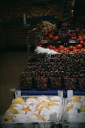 A variety of fresh produce is displayed in a supermarket setting. The foreground features trays of neatly wrapped yellow pears. Behind, there are containers filled with ripe red cherries and clusters of red tomatoes. The dim lighting gives a cozy, market feel.