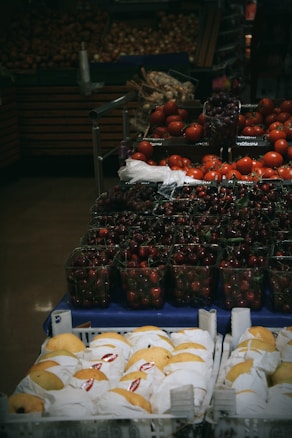 A variety of fresh produce is displayed in a supermarket setting. The foreground features trays of neatly wrapped yellow pears. Behind, there are containers filled with ripe red cherries and clusters of red tomatoes. The dim lighting gives a cozy, market feel.