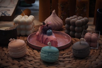 A collection of decorative candles is arranged on a woven mat. The candles are shaped like macarons, bubbles, and a cake topped with fruit. They come in soft pastel colors including pink, blue, and cream. The setting gives a cozy and artisanal feel, with a focus on aesthetically pleasing design.