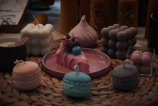 A collection of decorative candles is arranged on a woven mat. The candles are shaped like macarons, bubbles, and a cake topped with fruit. They come in soft pastel colors including pink, blue, and cream. The setting gives a cozy and artisanal feel, with a focus on aesthetically pleasing design.