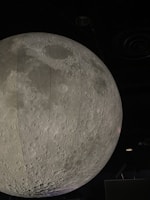A large, detailed model of the moon is suspended indoors, showcasing the lunar surface with its craters and texture. The setting is dimly lit, emphasizing the grey tones and realistic portrayal of the moon.