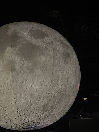 A large, detailed model of the moon is suspended indoors, showcasing the lunar surface with its craters and texture. The setting is dimly lit, emphasizing the grey tones and realistic portrayal of the moon.