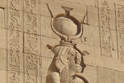 Ancient Egyptian wall carving featuring hieroglyphics and a figure with detailed headdress and attire. The stone surface appears weathered but well-preserved, showcasing intricate carvings and historical inscriptions.