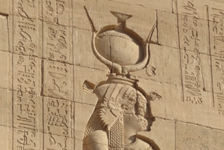 Ancient Egyptian wall carving featuring hieroglyphics and a figure with detailed headdress and attire. The stone surface appears weathered but well-preserved, showcasing intricate carvings and historical inscriptions.
