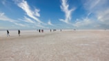 A vast, open salt flat extends to the horizon under a bright blue sky with scattered, wispy clouds. Several people are walking or standing, some appearing to be in groups, spread out across the expansive terrain.