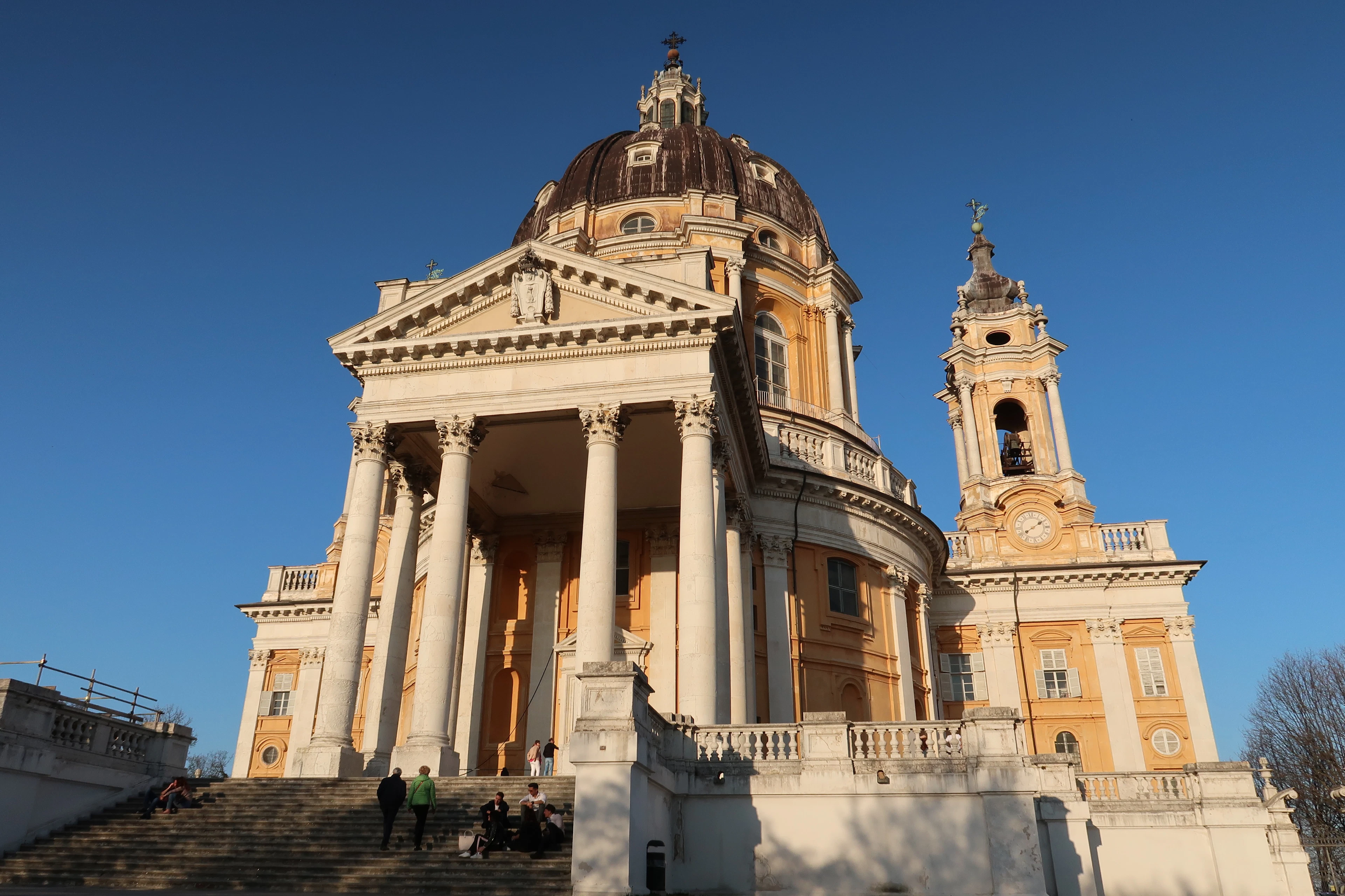 The golden stone facade, columned portico and dome of the Basilica di Superga on its hilltop above Turin, lit by low sun against a deep blue sky with visitors sitting on the steps below.