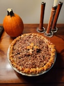 A freshly baked pie with a detailed crust sitting on a wooden table next to an orange pumpkin and a set of wooden candlesticks with partially melted candles.
