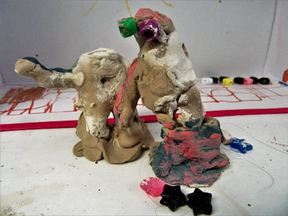 Two handmade clay sculptures with multicolored paint stand on a surface scattered with small star-shaped objects. The sculptures appear abstract, with rough, uneven surfaces and hints of green, pink, and blue paint. In the background, some markers or paint tubes lay on a white sheet, which has sketches or drawings in red and other colors.