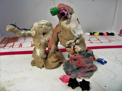 Two handmade clay sculptures with multicolored paint stand on a surface scattered with small star-shaped objects. The sculptures appear abstract, with rough, uneven surfaces and hints of green, pink, and blue paint. In the background, some markers or paint tubes lay on a white sheet, which has sketches or drawings in red and other colors.
