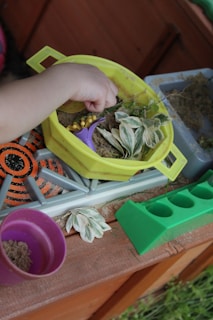 A child's hand is playing with a small kitchen playset. The playset includes a yellow bowl containing sand, a purple cup, some faux leaves, and small plastic yellow corn pieces. The setting appears to be outdoors, with a play kitchen counter visible.