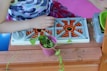A child wearing a colorful, fish-patterned shirt is playing with a toy stove. There is a pink flower pot with green plants inside it placed on a wooden surface. The toy stove features orange and black spiral patterns on its burners and is located next to a green bowl and sand scattered around.