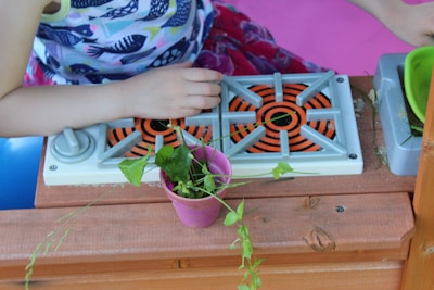 A child wearing a colorful, fish-patterned shirt is playing with a toy stove. There is a pink flower pot with green plants inside it placed on a wooden surface. The toy stove features orange and black spiral patterns on its burners and is located next to a green bowl and sand scattered around.