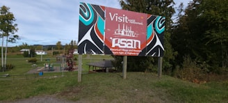 A colorful signboard with artistic patterns is placed in a grassy outdoor area. The sign features text promoting a historical village and museum, with stylized images in red, blue, black, and white. Behind the sign, there are trees, a playground, and buildings, suggesting a rural or suburban environment.
