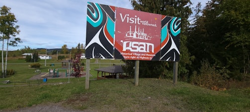 A colorful signboard with artistic patterns is placed in a grassy outdoor area. The sign features text promoting a historical village and museum, with stylized images in red, blue, black, and white. Behind the sign, there are trees, a playground, and buildings, suggesting a rural or suburban environment.