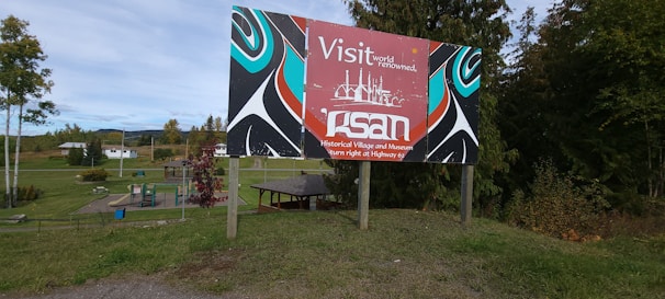 A colorful signboard with artistic patterns is placed in a grassy outdoor area. The sign features text promoting a historical village and museum, with stylized images in red, blue, black, and white. Behind the sign, there are trees, a playground, and buildings, suggesting a rural or suburban environment.