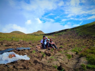 A group of people sitting on a hillside field with patches of green grass and earth. A heap of harvested potatoes lies on a tarp in the foreground. The sky is a vibrant mix of blue with scattered clouds, and the landscape has rolling hills in the background.
