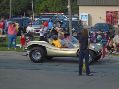 A street scene featuring a small gold dune buggy with a driver and child sitting inside. People, including children and adults, are gathered on the sidewalk and appear to be watching or participating in an event. A red balloon and blue balloon are held by one of the children. Various parked cars and a building with a movie rental kiosk are in the background.