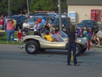 A street scene featuring a small gold dune buggy with a driver and child sitting inside. People, including children and adults, are gathered on the sidewalk and appear to be watching or participating in an event. A red balloon and blue balloon are held by one of the children. Various parked cars and a building with a movie rental kiosk are in the background.